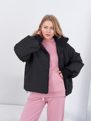 Woman poses in a relaxed stance, wearing a black puffer jacket over a pink hoodie and matching...
