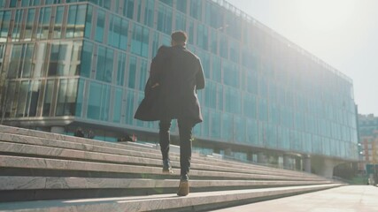 Cinematic shot in urbanscape, man running up of upstairs in business district. Back view of stylish african male person in black coat moving to office building, move to future success, career growth