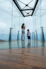 A woman and a child are walking on a bridge. The woman is holding a cell phone. The bridge is made of glass and wood