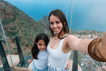 A woman and a child are taking a selfie from a high vantage point. The woman is wearing a white tank top and the child is wearing a white shirt. The photo has a happy and lighthearted mood