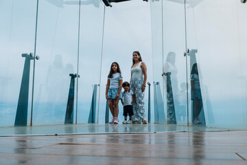 A woman and two children stand in front of a glass wall. The woman is wearing a white shirt and the children are wearing white