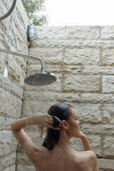 woman showering outdoors under rain showerhead with stone wall background, natural light, relaxing moment, fresh water spa