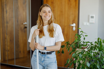 A joyful woman with long hair stands in a stylish interior, holding a cleaning tool, embodying...