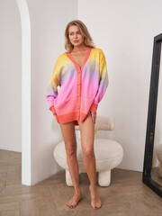 Woman stands indoors wearing a large, vibrant cardigan with orange, pink, and yellow gradient,...