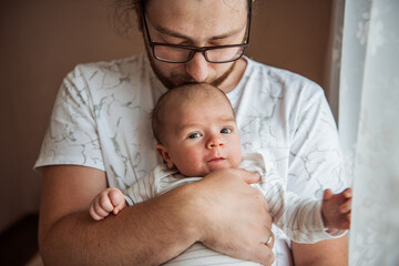Tender moment as a father cradles his newborn baby near a window. The soft lighting highlights the bond and warmth shared between them, capturing the essence of parenthood
