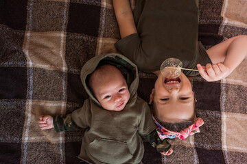 Older sister lying upside down on a checkered blanket, enjoying a sweet isomalt lollipop while bonding with her newborn baby brother, creating a cozy moment of family joy and love