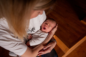 Young mother cradling her yawning newborn baby in her arms during a quiet moment at home. Natural indoor lighting creates an intimate atmosphere while the infant displays signs of tiredness