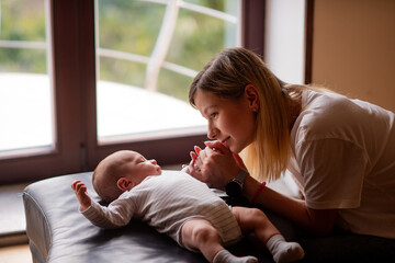 Young mother holding her newborn's hands while lying on a cozy sofa in front of a window, embracing the joys of motherhood and cherishing the bond of childcare