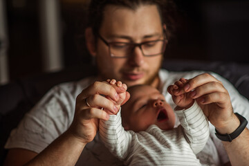 Father lovingly holds his newborn baby's hands, gently lifting them while the baby yawns. The intimate moment captures the warmth and connection of early parenthood in a cozy home setting