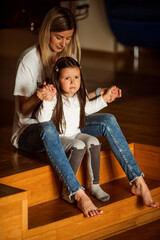 Young mother sitting on wooden stairs at home, holding hands with her sad little daughter, providing comfort and expressing unwavering support during a challenging moment