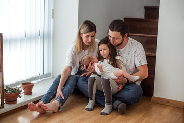 Young family of four sitting on a wooden floor near stairs, sharing a tender moment. Parents gently hold their newborn while the older sister looks on with curiosity and affection