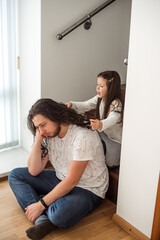 Young girl is playfully braiding her father's long hair as he sits on a staircase. The scene captures a tender family moment, highlighting the bond and joy of spending time together at home
