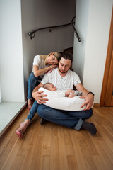 Young mother and father sitting on the floor beneath the stairs, cradling their sleeping newborn baby in a cozy baby nest, embodying love and tenderness in their serene home environment