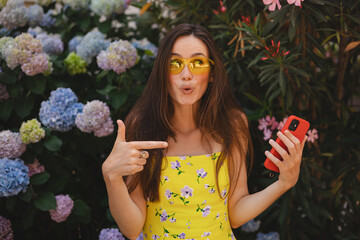 Yes, great news. Portrait of excited woman celebrating win, using mobile phone and pointing on gadget, smiling young lady having success, holding cellphone, she wear yellow dress and sunglasses.
