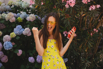 Happy joyful woman winner excited lady in city hold mobile phone gadget addict amazing news victory shock surprise celebrate success online money win prize outdoors near blossom hydrangea.