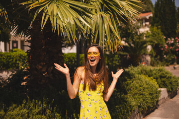Brunette long hair tourist woman wear yellow dress and sunglasses standing near palm tree, enjoy of trip vacation. Happy girl on vacation smiling, walking. Hot summer day mood. Here i am, wow emotions