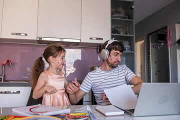 Father wearing headphones, focusing on his laptop while his young daughter plays nearby, frequently interrupting him at the kitchen table, balancing work and parenting challenges