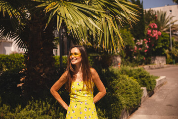Brunette long hair tourist woman wear yellow dress and sunglasses standing near palm tree, enjoy of trip vacation. Happy girl on vacation smiling, walking. Hot summer day mood.