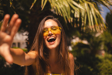 Playful young woman enjoying a sunny summer day in a tropical setting. Smiling, wearing yellow sunglasses, dress reaching hand toward the camera, wave hand hi. Vibrant lifestyle and joyful emotions. © zvkate