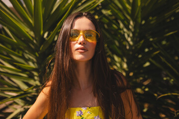 Caucasian woman look serious travel vacations for visiting nature environment, beautiful female tourist with brunette hair posing near palm leaves. Girl wear yellow sunglasses and floral print dress. 