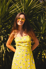 Smiling young woman standing in front of tropical palm tree. Wearing yellow sunglasses and a floral...