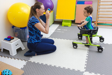 Handsome young boy with movement disorders practices walking with support from a therapist in rehabilitation clinic