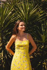 Smiling young woman look at side standing in front of tropical palm tree. Wearing yellow floral...