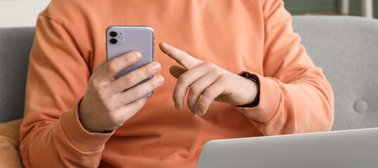 Young man with laptop using mobile phone on sofa at home, closeup