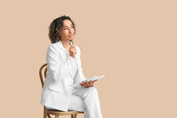 Mature thoughtful businesswoman with pen and paper sheet sitting on chair on beige background