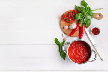 Cooking pot of tomato sauce with basil, garlic and chili peppers on white wooden background