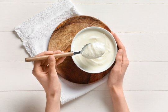 Female hands with bowl of tasty yogurt and spoon on white wooden background