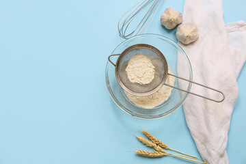 Metal sieve with flour in bowl, whisk, garlic and napkin on color background