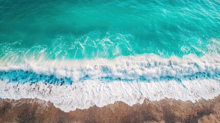 Fototapeta premium Aerial view of a serene turquoise ocean meeting a sandy shore with a frothy white wave breaking onto the beach. The water transitions from deep turquoise to light aqua, creating a beautiful gradient. 