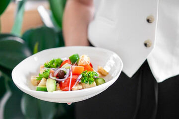A waiter in a white uniform presents a fresh salad with tomatoes, cucumbers, olives, red onions,...