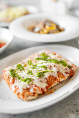 A close-up of a rectangular pizza slice with melted cheese, tomato sauce, and green pesto on a white plate. The background is blurred, creating a warm dining atmosphere.