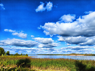 Obraz premium Vast blue sky with dynamic clouds reflecting over serene lake and lush greenery during daylight