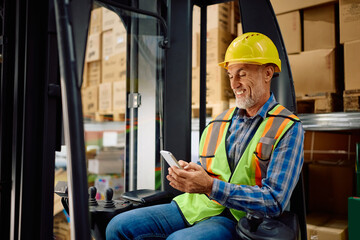 Happy warehouse worker texting on cell phone while sitting in forklift at storage compartment.