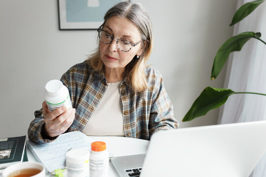 Aged woman holding bottle of dietary supplement and reading label with instruction, ingredients or brand name carefully before ordering vitamins online using laptop and web application