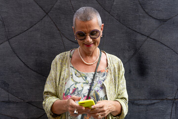 elegant smiling mature woman with shaved grey hair texting on cell phone in front of black wall