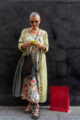 elegant smiling mature woman with shaved grey hair texting on cell phone in front of black wall