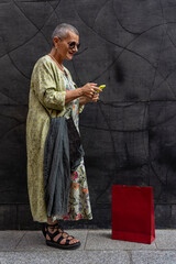 elegant smiling mature woman with shaved grey hair texting on cell phone in front of black wall
