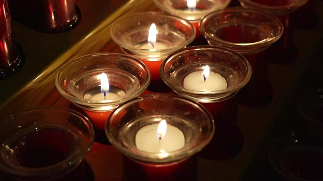 Group of burning votive candles in red and transparent glass holders on a wooden shelf in a church