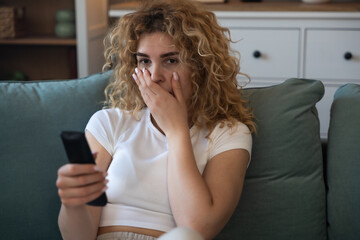 Young woman sitting on couch at home holding remote control, shocked and heartbroken while watching...