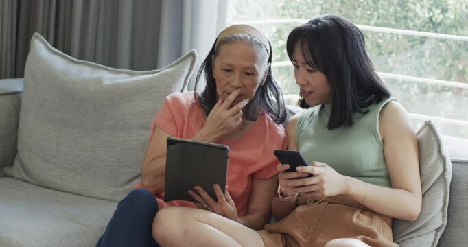 Chinese Asian mother and daughter sharing pictures loading phone and browsing tablet on gray sofa