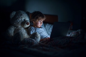 A young boy is sitting on a bed with a teddy bear next to him. He is looking at a laptop computer
