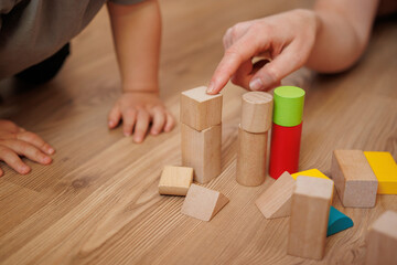 Close-up of adult hand pointing at wooden geometric blocks while a toddler learns to build. Educational activity for preschool children at home.