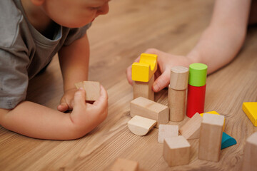 Boy plays with his mother with wooden blocks sitting on the floor at home. Educational activity for developing fine motor skills.