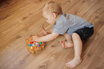 Baby boy playing with wooden fishing toy while sitting on floor at home. Educational activity for fine motor skills development.
