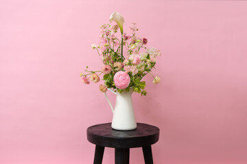 Soft pastel flowers in a white ceramic jug placed on a dark wooden stool, styled with a pink studio background.