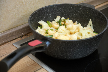 Fresh cauliflower florets and green beans are being cooked in a non-stick frying pan on an electric stove for a healthy dish.
